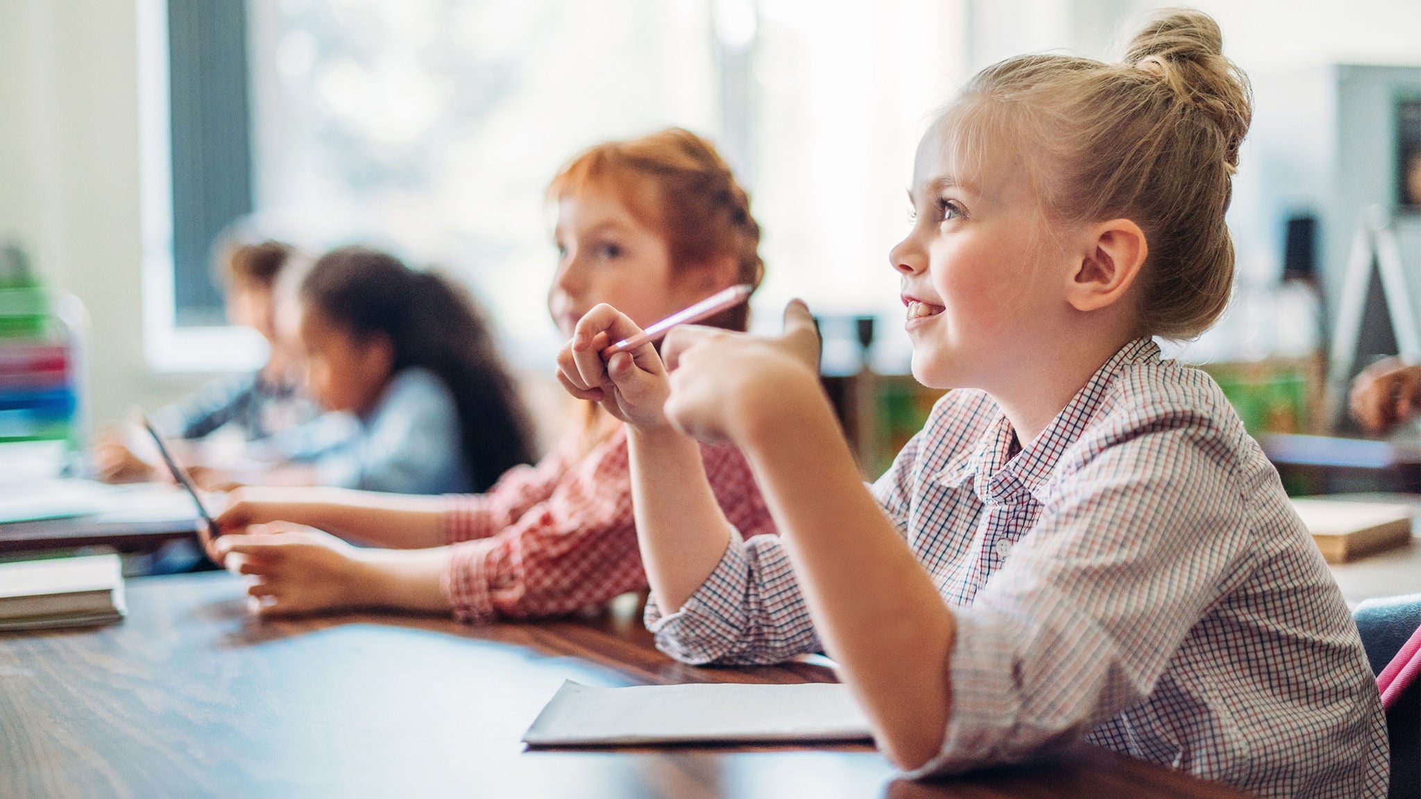 A group of young students sits at desks in a brightly lit classroom, focusing on their schoolwork