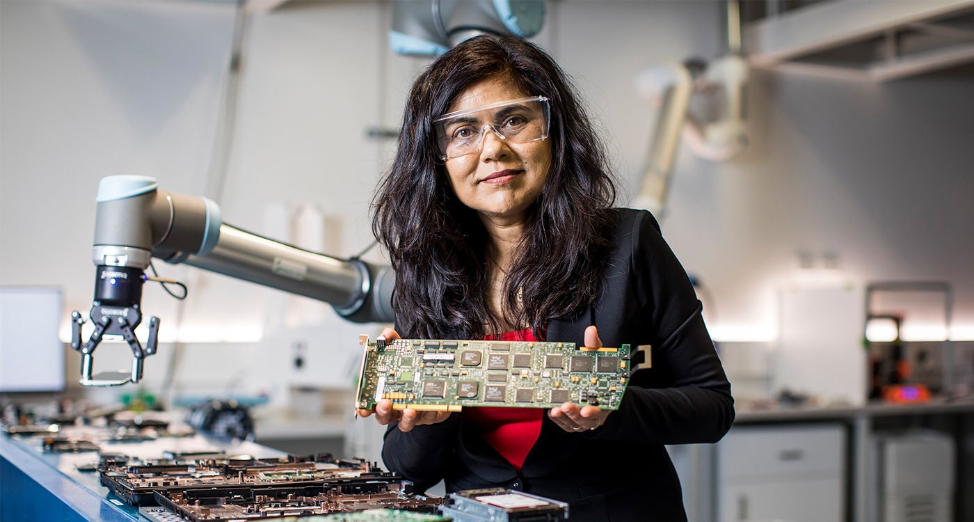 An olive-skinned woman in a red top, black blazer and protective eyewear standing in a lab and holding a circuit board. 