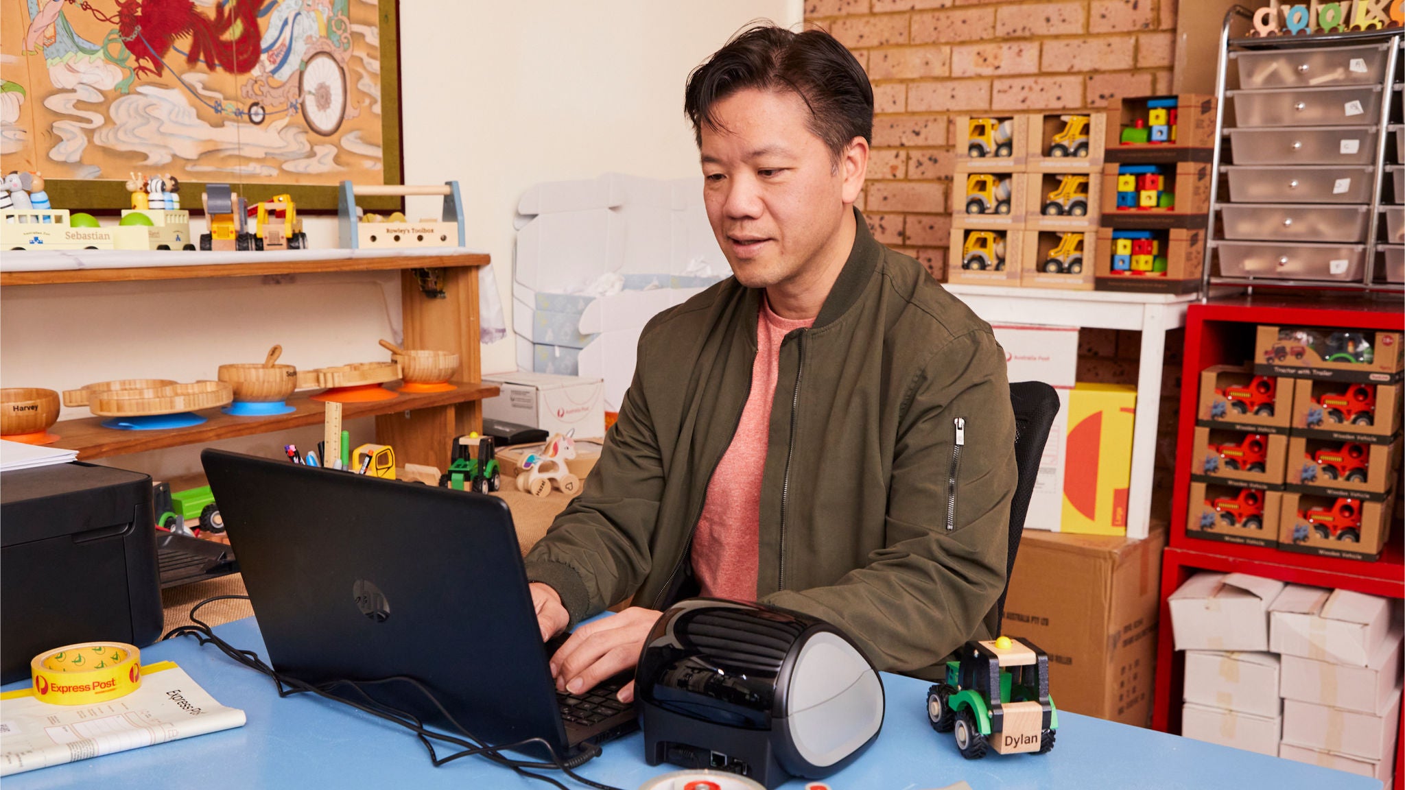 An male sits at a blue desk using a laptop, surrounded by various office and toy item.
