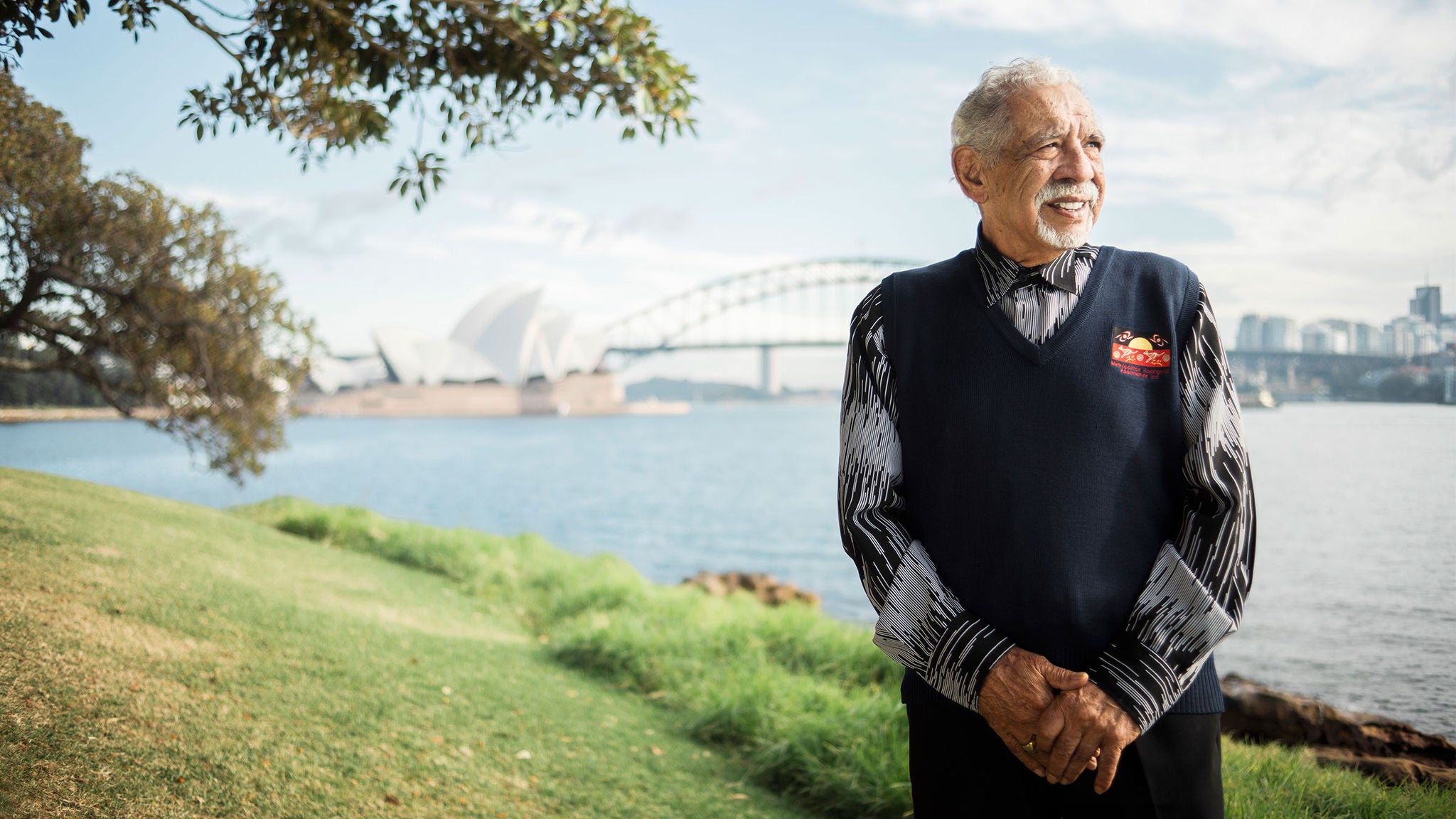 Gadigal Elder, Uncle Allen Madden, smiles at camera on Gadigal land with the Sydney Harbour Bridge in the distance behind him.