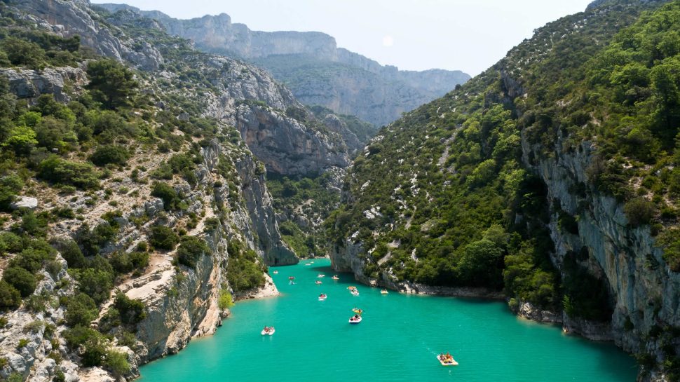 A panoramic view of the Verdon Gorge in France. Azure, still water runs through the middle of this scene, subtly winding through the rock formations that surround it. Huge rocks forming the gorge stand tall to both the left and right. There’s dense shrubbery forming on the rocks. People can be seen kayaking through the crystal blue water. 